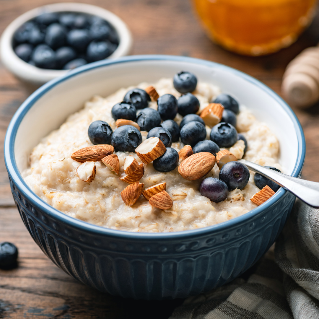 A blue bowl filled with oatmeal topped with fresh blueberries, sliced almonds, and a few nuts, set on a rustic wooden table with a small bowl of blueberries and a jar of honey in the background.