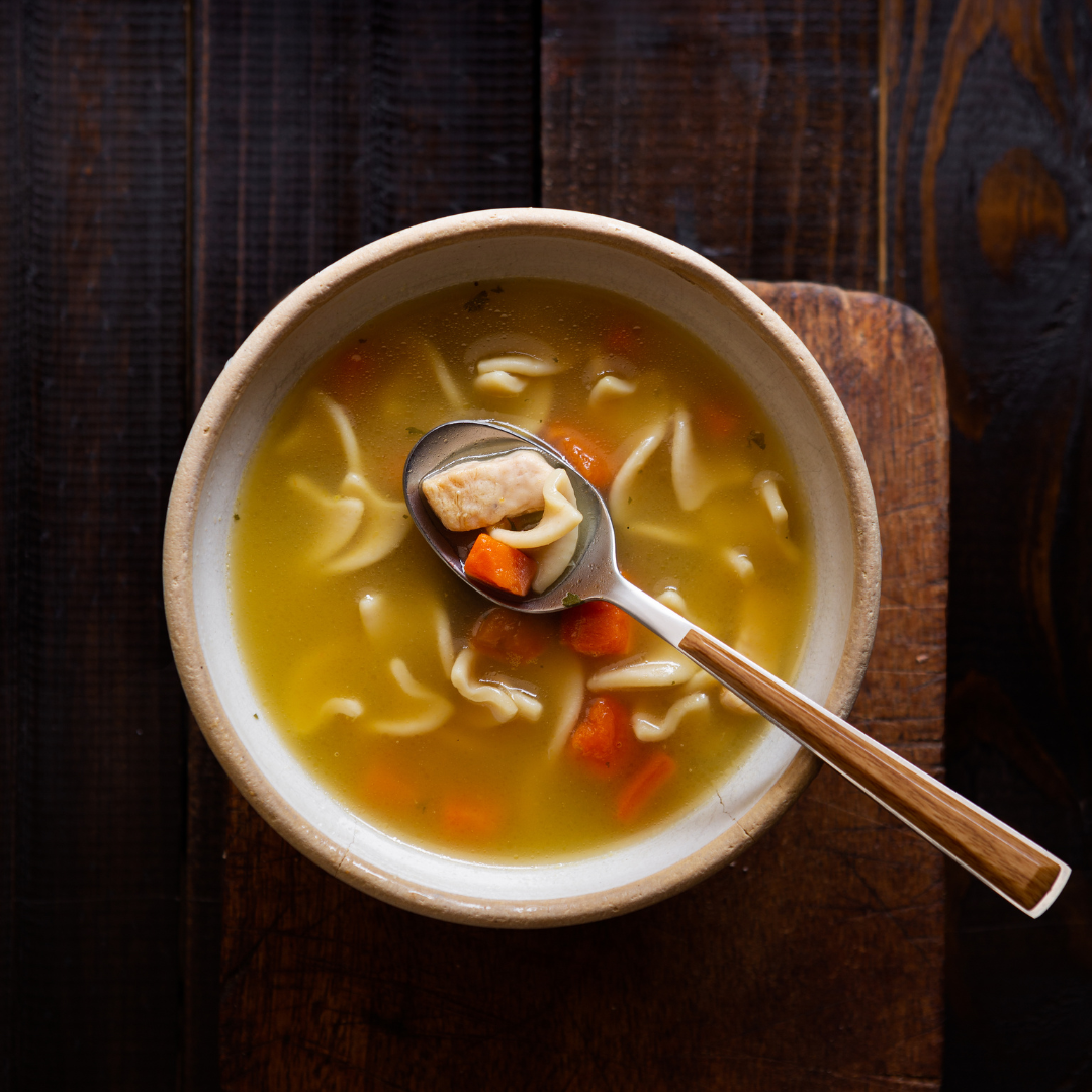 A top-down view of a ceramic bowl filled with chicken noodle soup, featuring egg noodles, carrots, and chunks of chicken, with a spoon resting in the bowl on a wooden board over a dark wood background.