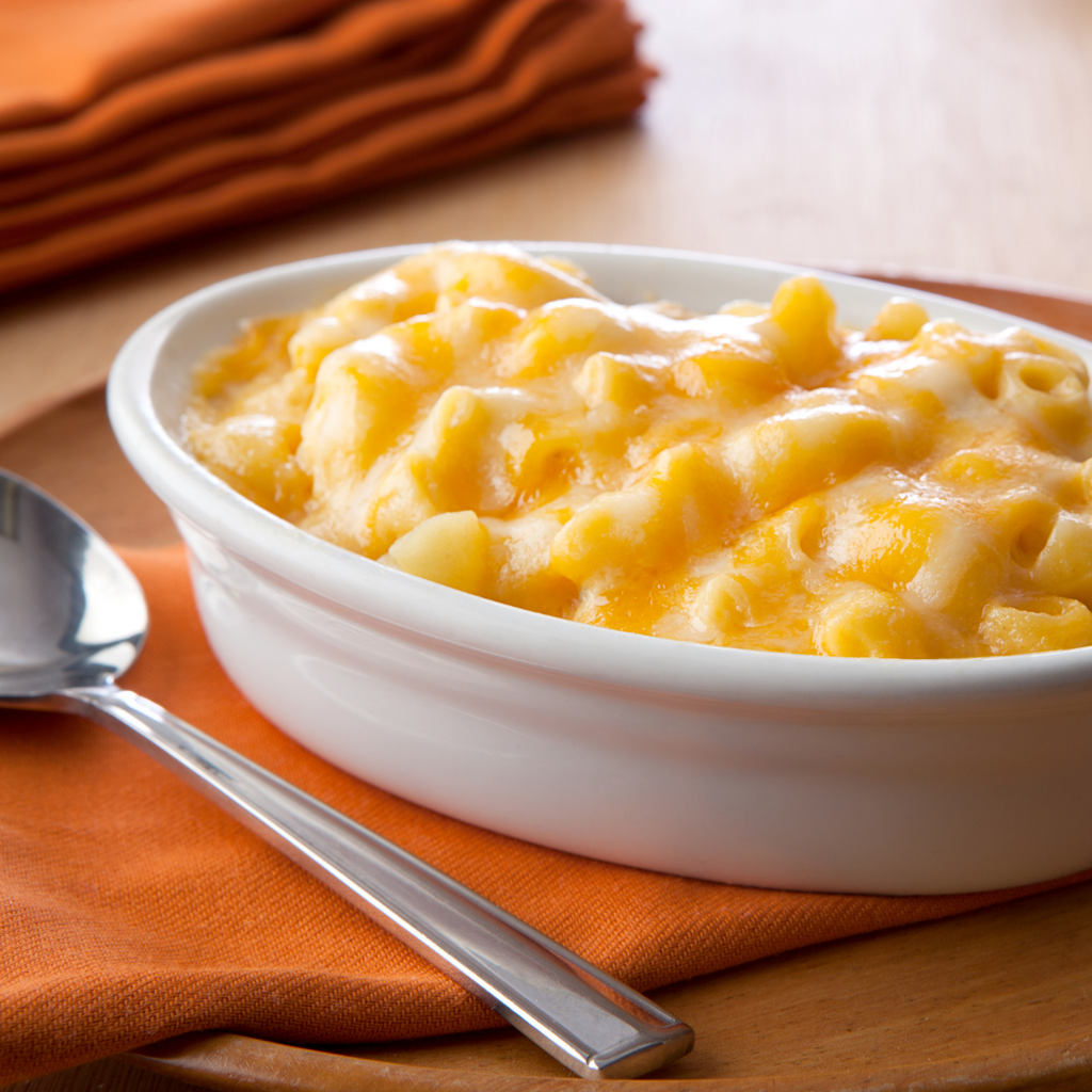 Mashed macaroni cheese in a white oval baking dish, served on an orange cloth napkin with a metal spoon on a wooden table.