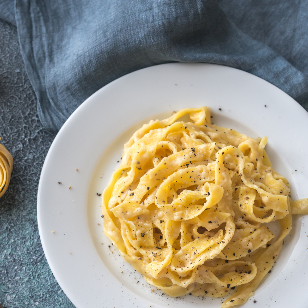 A top-down view of creamy fettuccine pasta on a white plate, sprinkled with black pepper, set against a textured slate surface with a blue linen napkin.