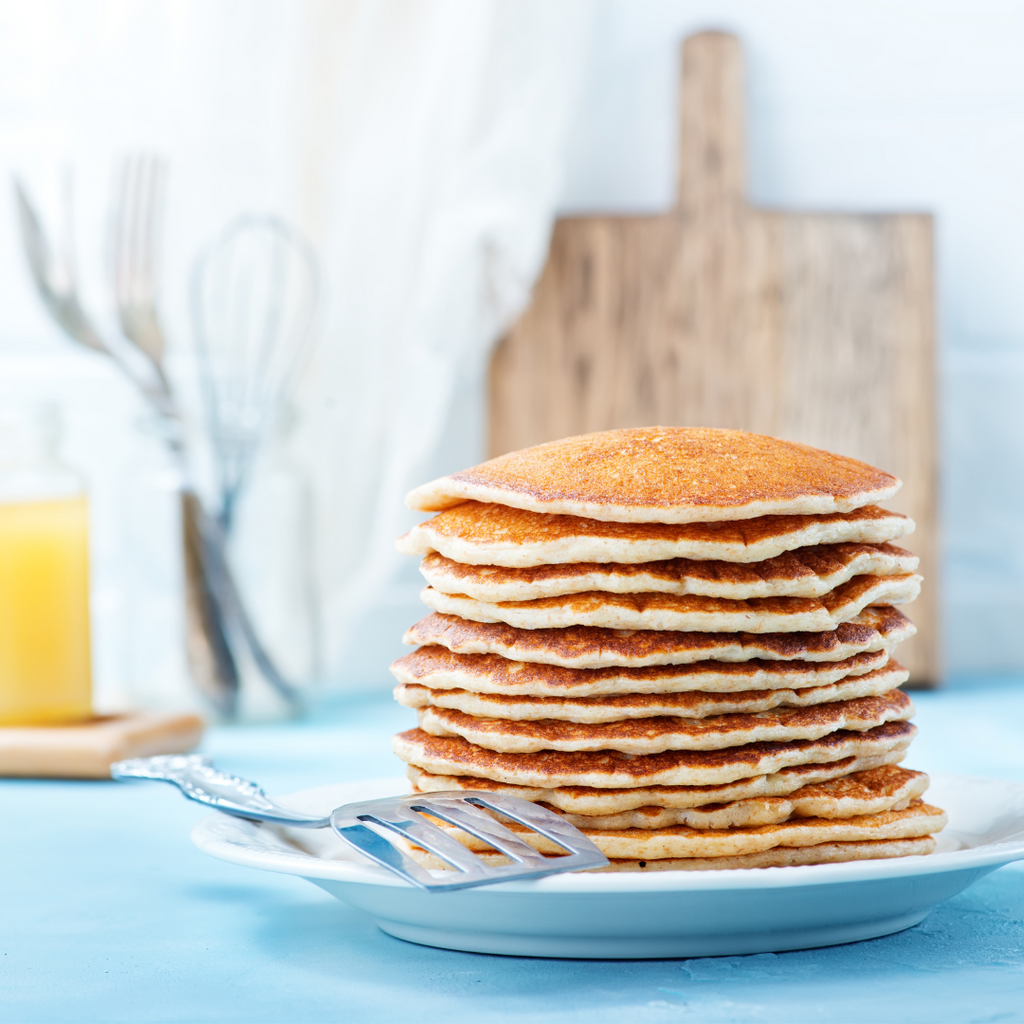A tall stack of golden-brown pancakes on a white plate with a metal spatula, set on a light blue surface with a wooden cutting board, whisk, and bottle in the softly blurred background.
