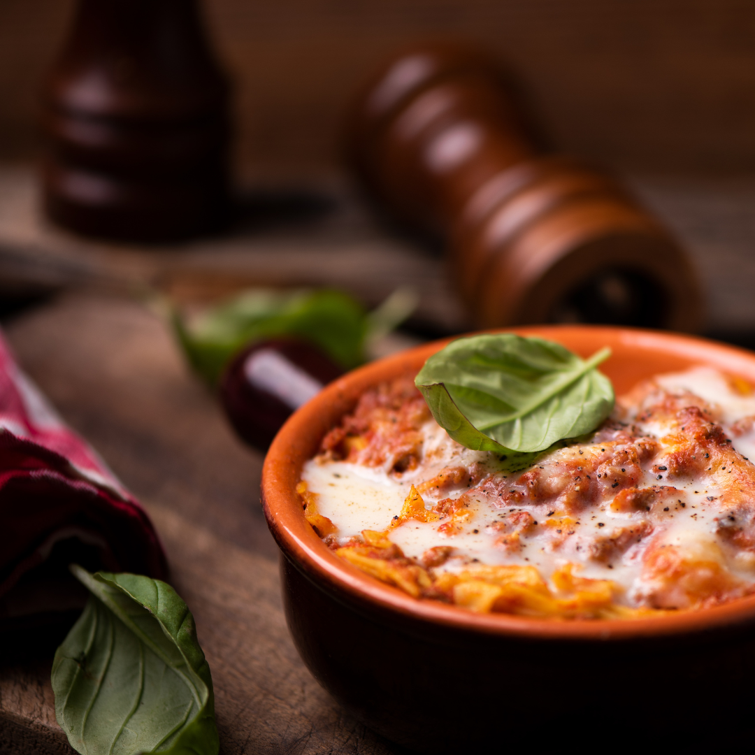 A close-up of a baked pasta dish in a small terracotta bowl, topped with melted cheese and a fresh basil leaf, set on a rustic wooden surface with blurred peppercorns and additional basil in the background.