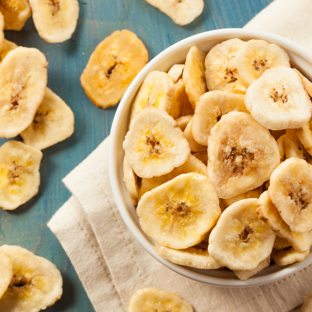 Banana chips in a white bowl on a beige napkin, with additional banana slices scattered on a blue wooden surface.