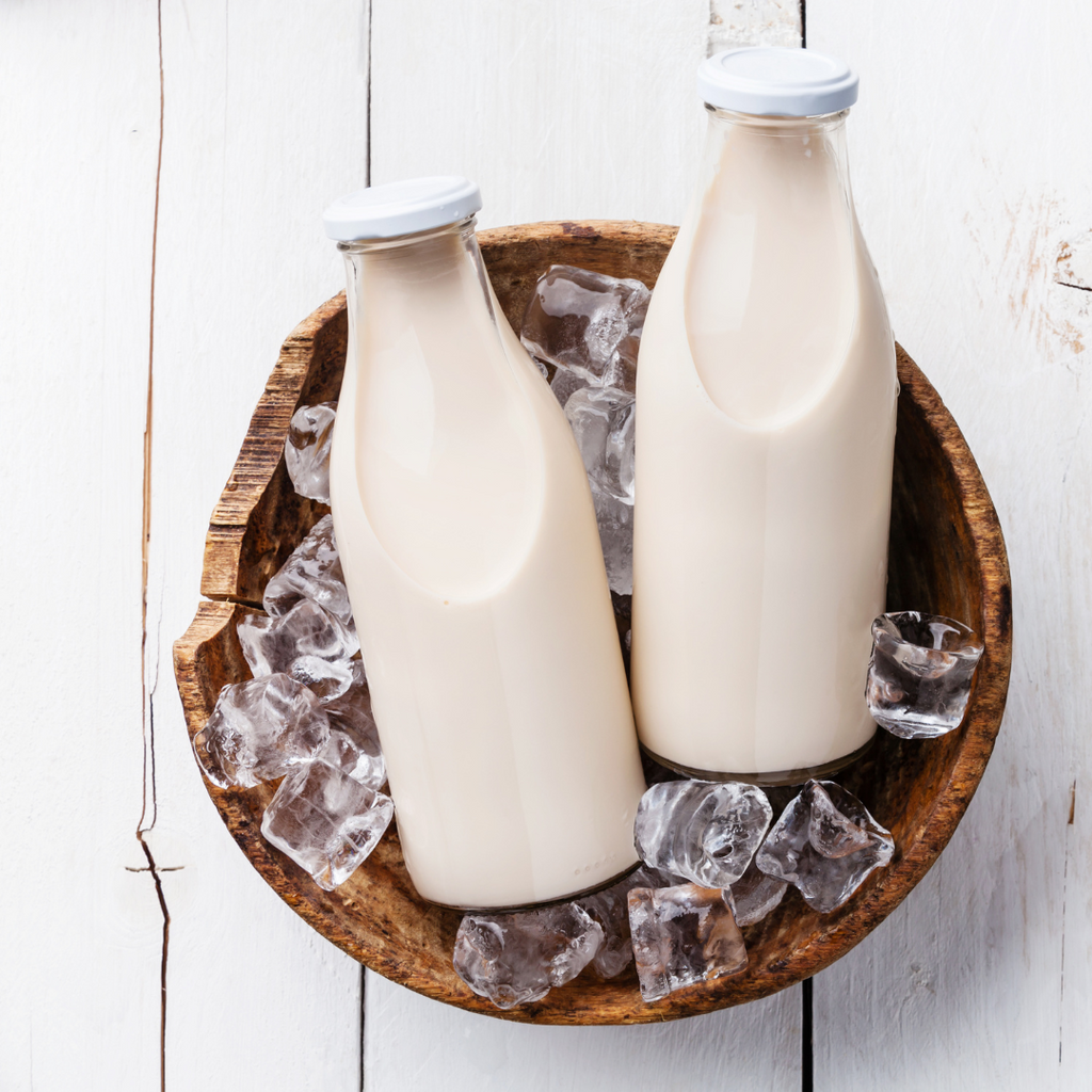 Two glass milk bottles filled with an off-white liquid, sealed with white caps, and arranged in a wooden bowl filled with ice cubes on a white wooden surface.