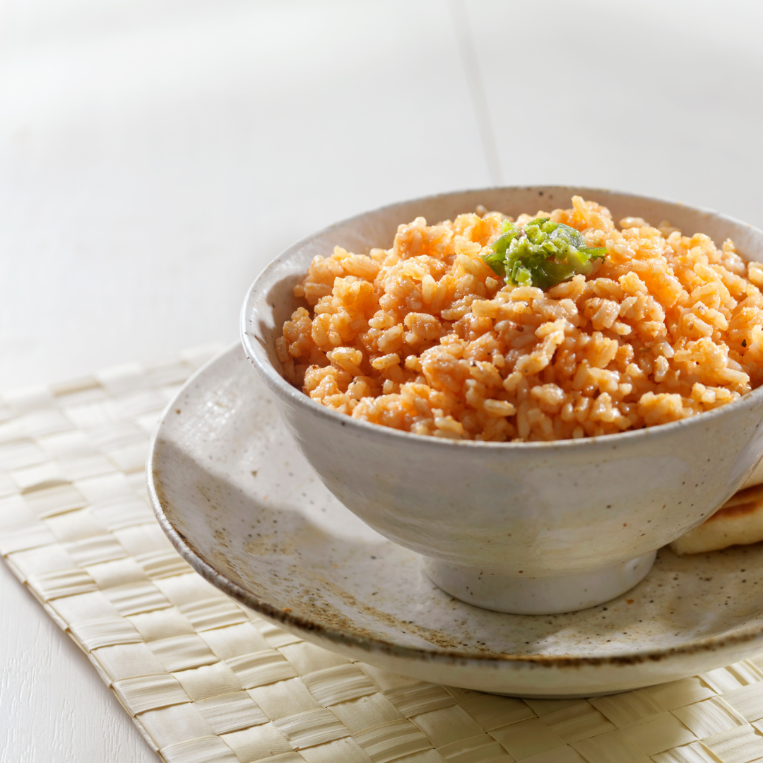 A ceramic bowl filled with orange-colored rice, garnished with a small green herb, set on a speckled ceramic plate over a woven placemat.