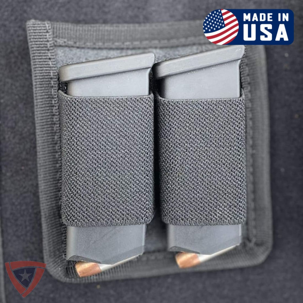 A close-up of a dark gray fabric dual magazine pouch holding two gray magazines with visible copper bullets, on a dark gray background. A 'Made in USA' badge appears in the top right, and a small shield logo appears in the bottom left.