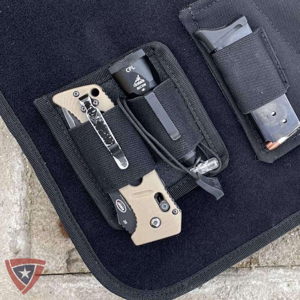 A black nylon knife and accessory organizer pouch laid on a dark surface, holding a tan folding knife, a black flashlight, and a magazine in a black holder. A small logo watermark appears in the lower-left corner.