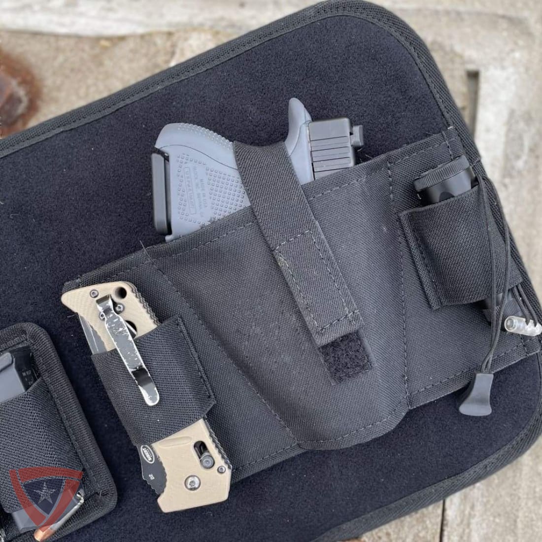 A close-up of a black nylon gun holster pouch containing a handgun, shown resting on a black zippered case outdoors on a stone surface.