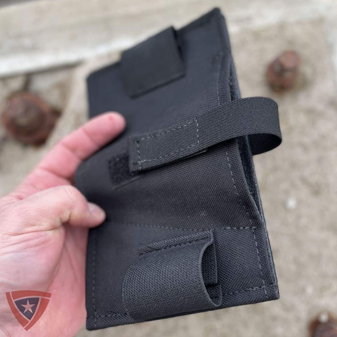 A black tactical pouch or holster made of durable fabric, held in a hand against a sandy outdoor background.