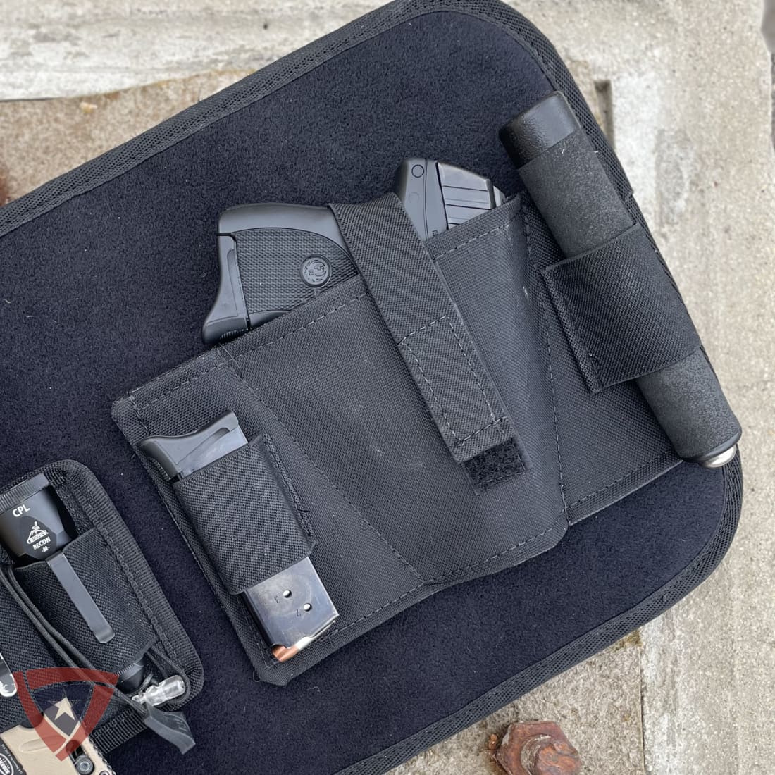 A black nylon holster organizer laid on a light stone surface, holding a handgun, magazines, and a cylindrical accessory. Part of another holster and a handgun are visible at the lower left.