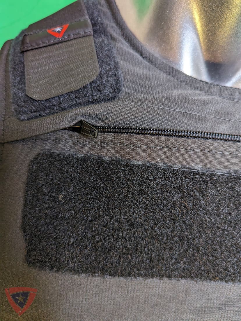 Close-up of a tactical vest or vest panel in olive drab and black, showing a hook-and-loop patch, a zipper, and a red triangular logo patch.