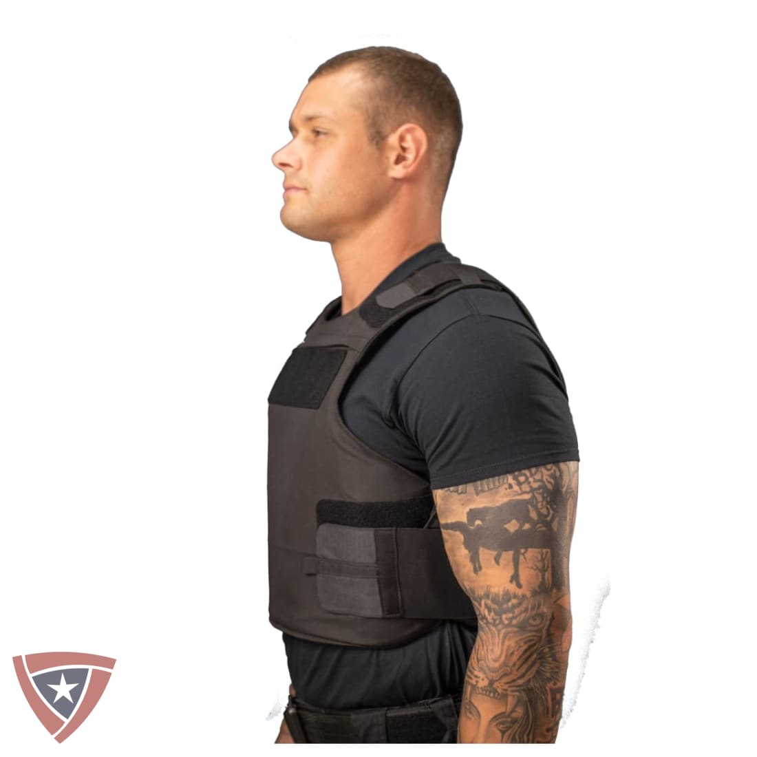 A man in profile wearing a dark gray tactical vest over a black t-shirt, shown against a plain white background with a small shield logo in the bottom-left corner.