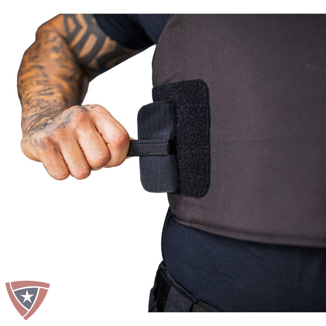 A close-up of a person's tattooed arm pulling a black strap off a dark gray tactical vest or body armor panel, with a small shield logo in the bottom-left corner.