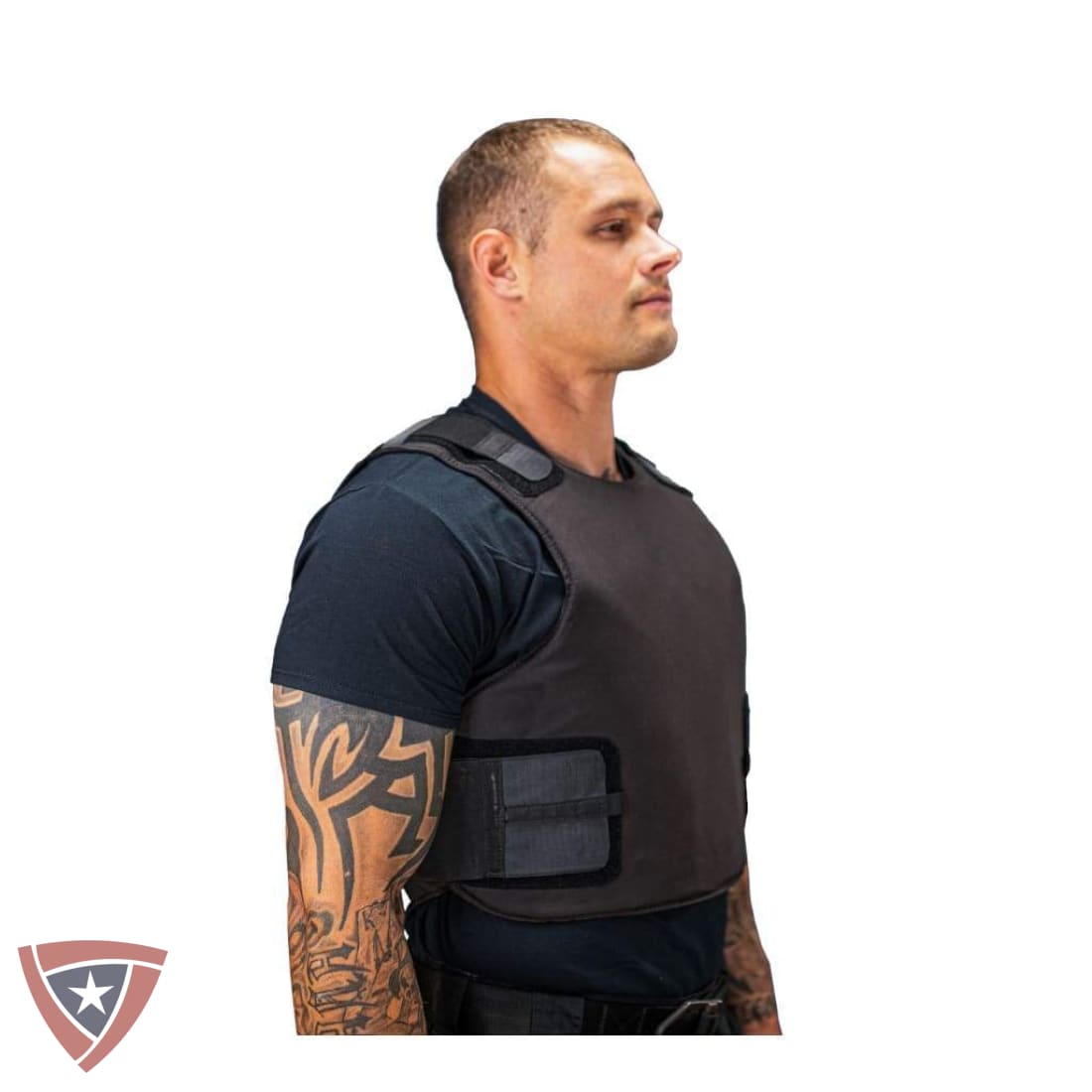 A man in profile wearing a dark gray tactical vest over a black t-shirt, shown against a plain white background with a small shield logo in the bottom-left corner.