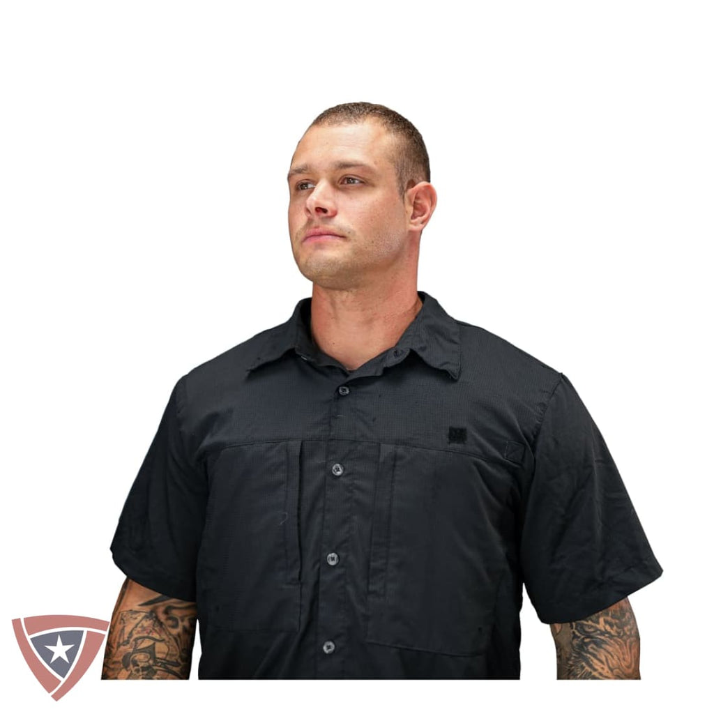 A man wearing a black short-sleeve button-up shirt with chest pockets, shown from the waist up against a plain white background. A small shield logo is visible on the left chest pocket, and a larger shield logo watermark appears in the bottom-left corner.