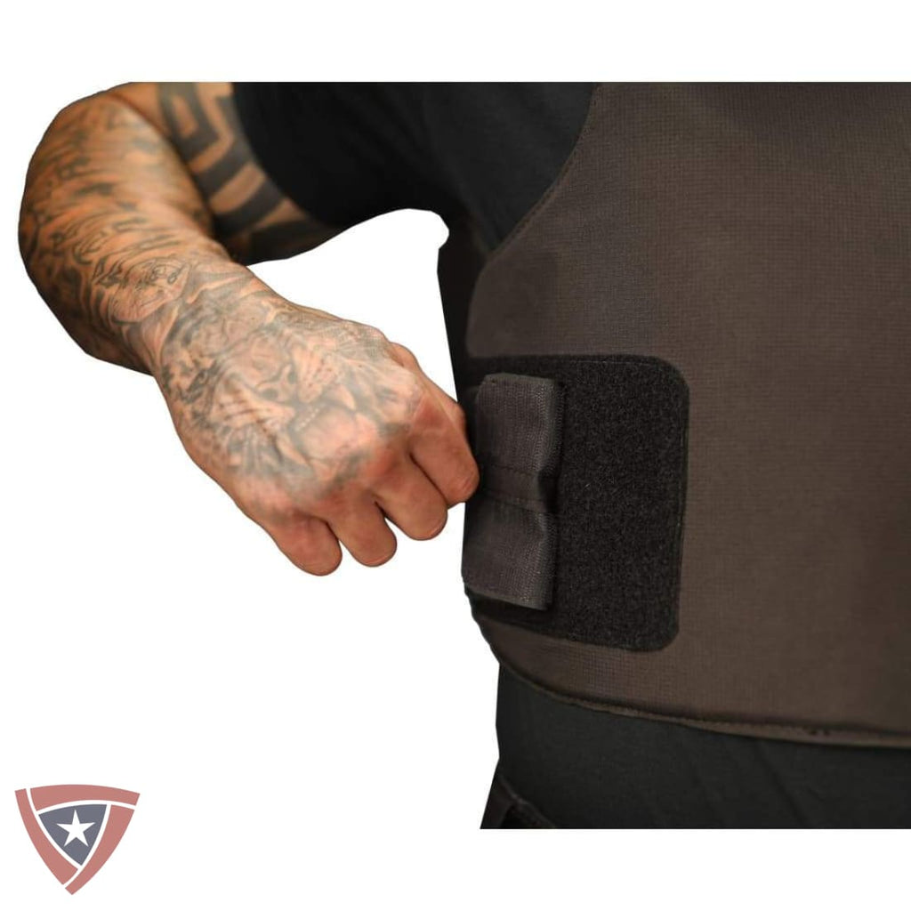 Close-up of a person's tattooed arm fastening a black hook-and-loop strap onto a dark brown tactical vest or body armor panel, shown against a white background.