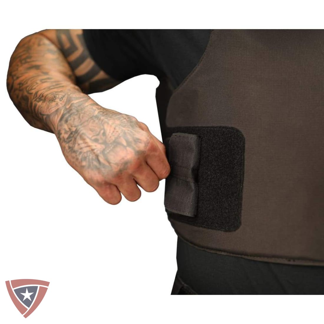 Close-up of a person's tattooed arm fastening a black hook-and-loop strap onto a dark brown tactical vest or body armor panel, shown against a white background.