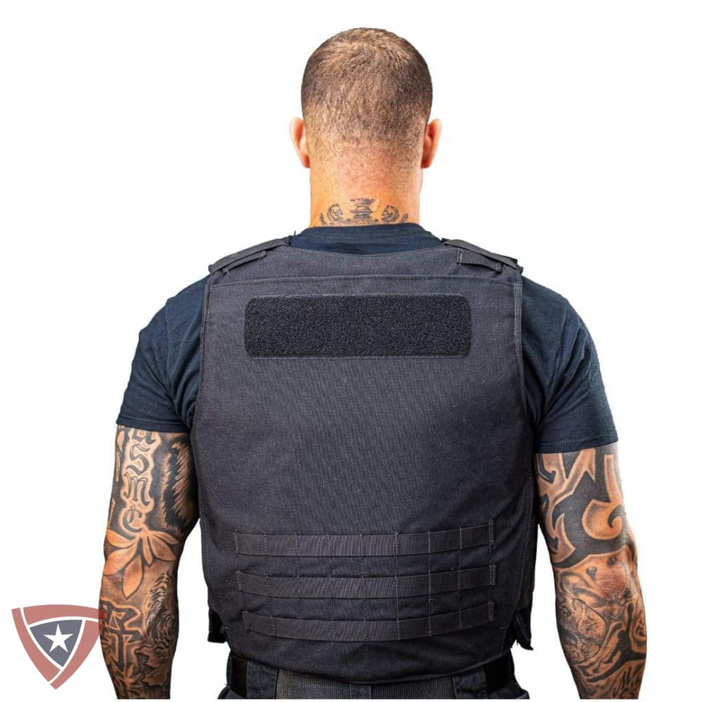A man viewed from the back wearing a black tactical vest over a dark blue t-shirt, against a plain white background.