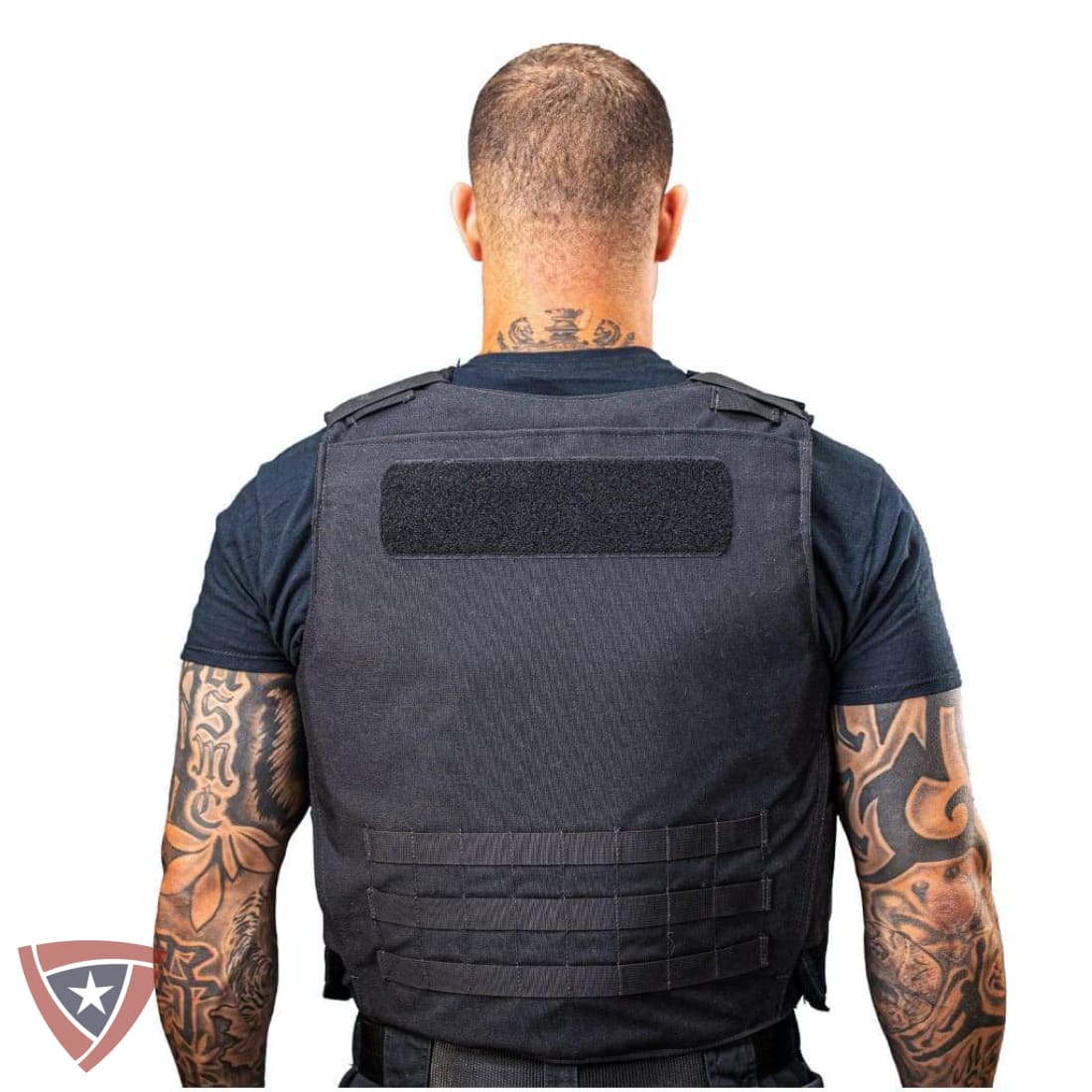 A man viewed from the back wearing a black tactical vest over a dark blue t-shirt, against a plain white background.