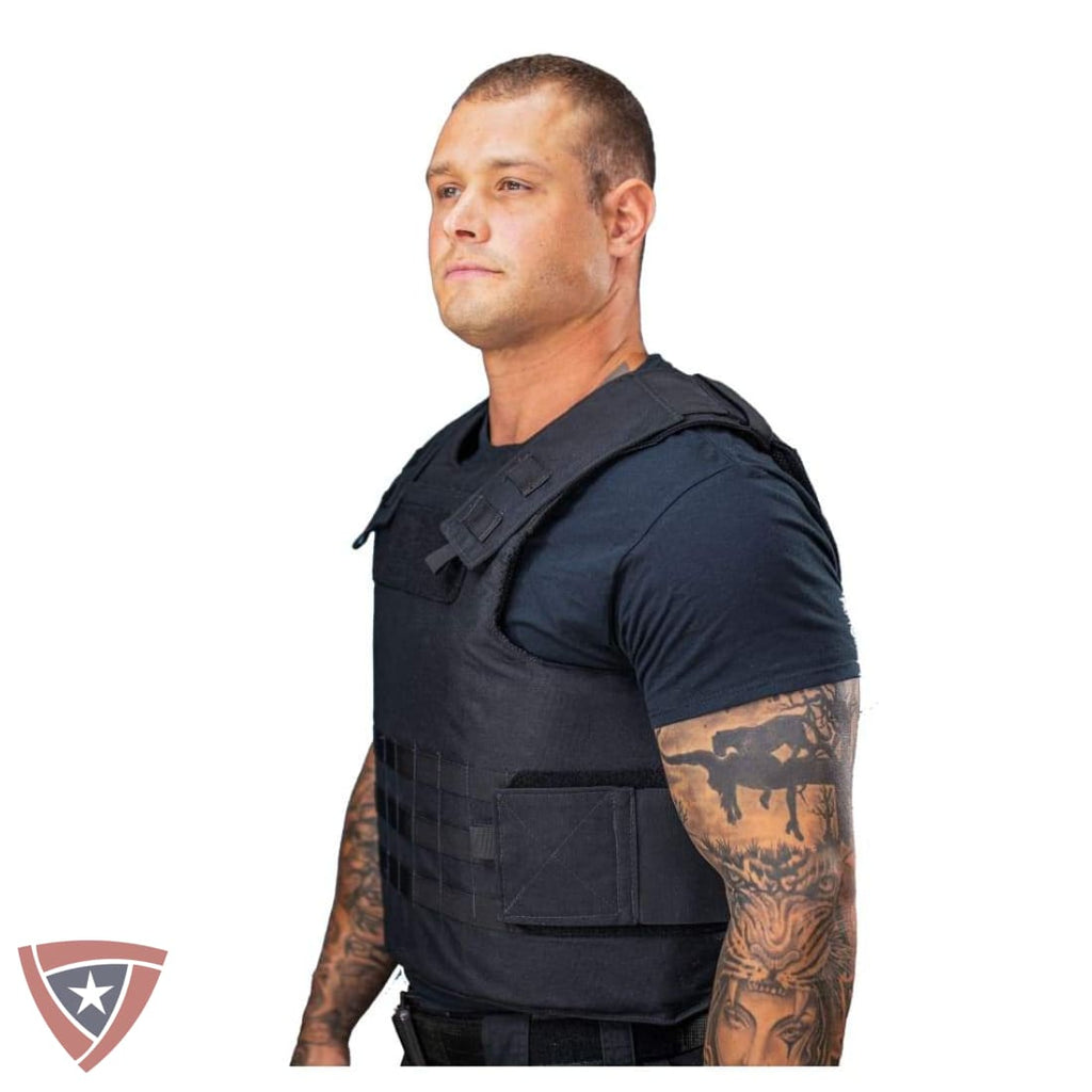 A man wearing a black tactical vest over a dark t-shirt, shown from the waist up against a plain white background. A shield-shaped logo is visible in the bottom-left corner.