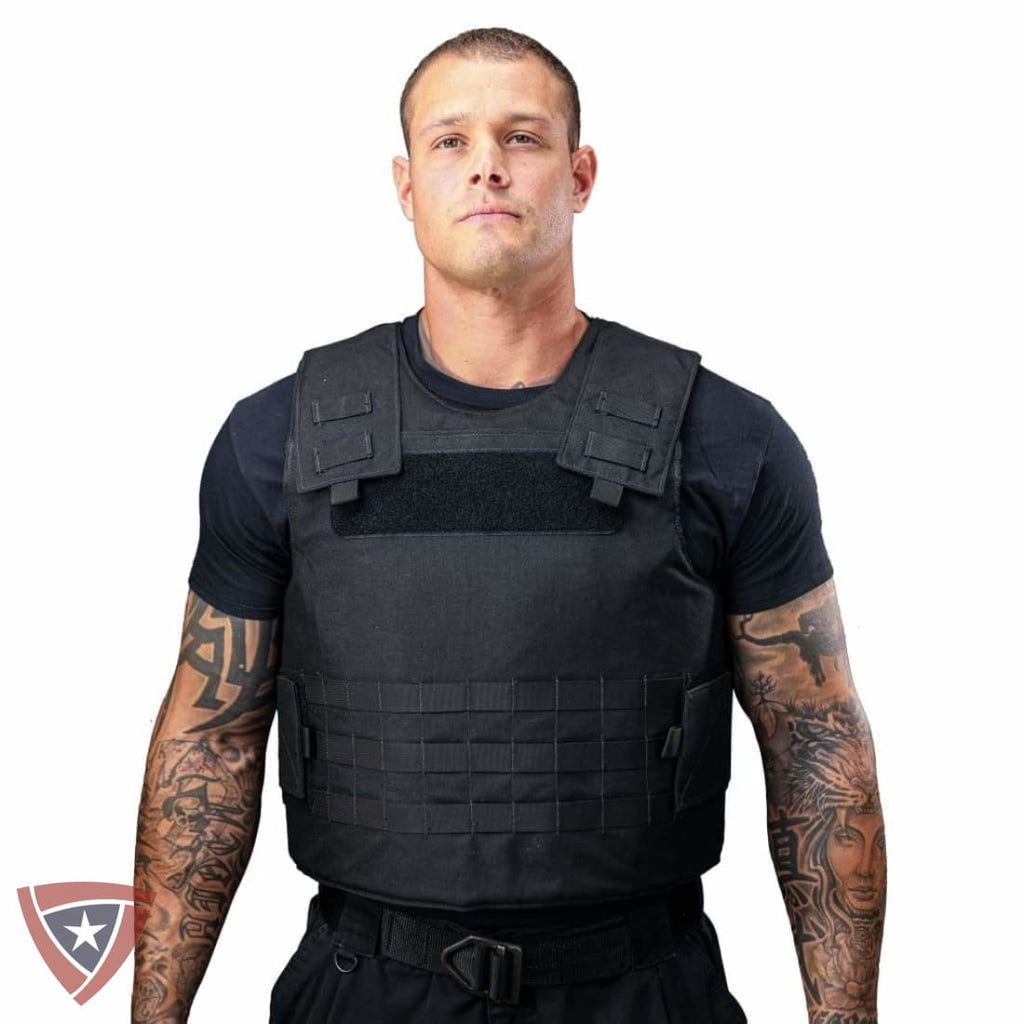 A man with tattooed arms wearing a black tactical plate carrier vest over a black t-shirt, shown against a plain white background.