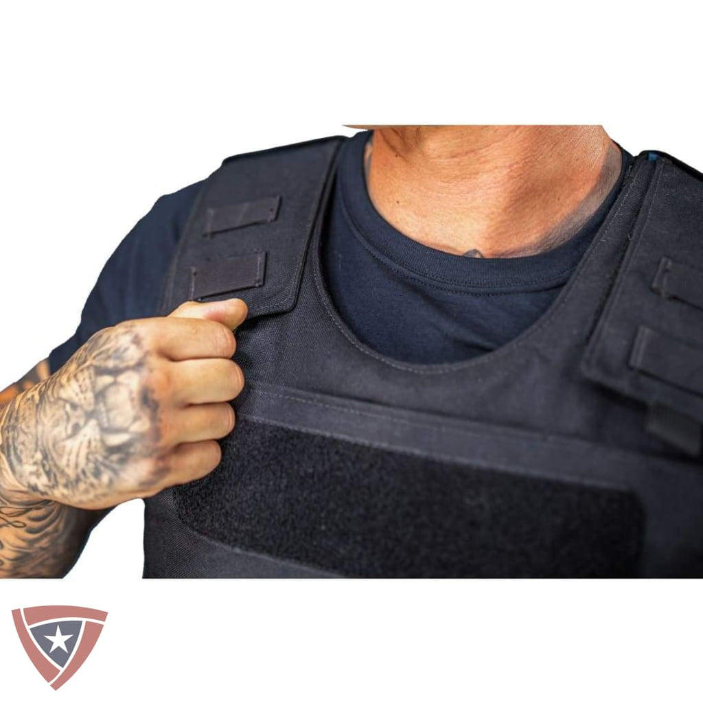 A close-up of a person wearing a black tactical plate carrier vest over a dark blue t-shirt, with a tattooed hand adjusting the shoulder strap. A shield-shaped logo is visible in the bottom-left corner.